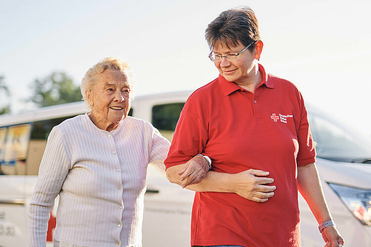 Eine ältere Dame hat sich bei einer DRK-Arbeiterin am Arm untergehakt und die beiden gehen glücklich bei schönem Wetter vor einem weißen DRK-Fahrzeug entlang.