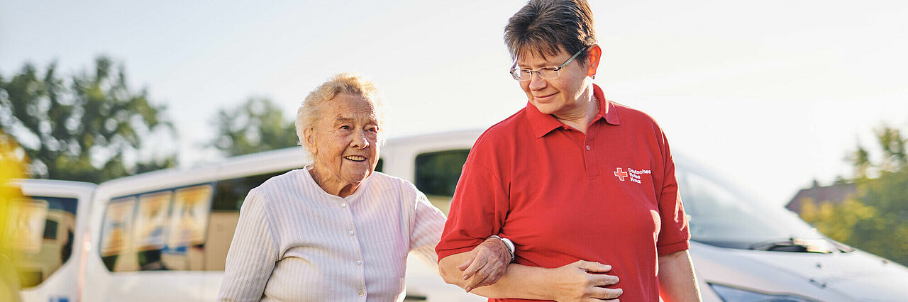 Eine ältere Dame hat sich bei einer DRK-Arbeiterin am Arm untergehakt und die beiden gehen glücklich bei schönem Wetter vor einem weißen DRK-Fahrzeug entlang.